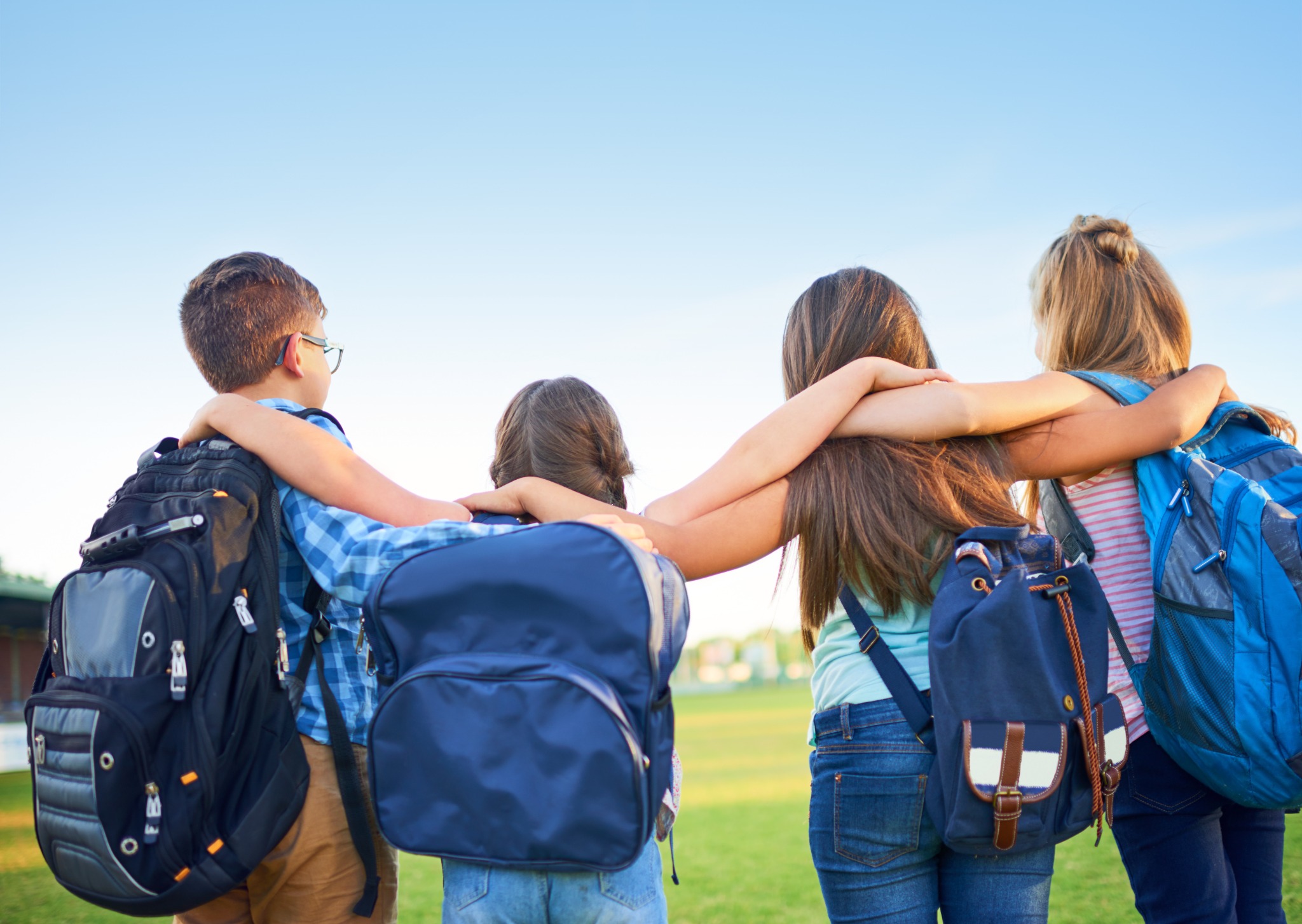 Vier Kinder mit Rucksäcken stehen im Freien und legen einander die Arme um die Schultern.
